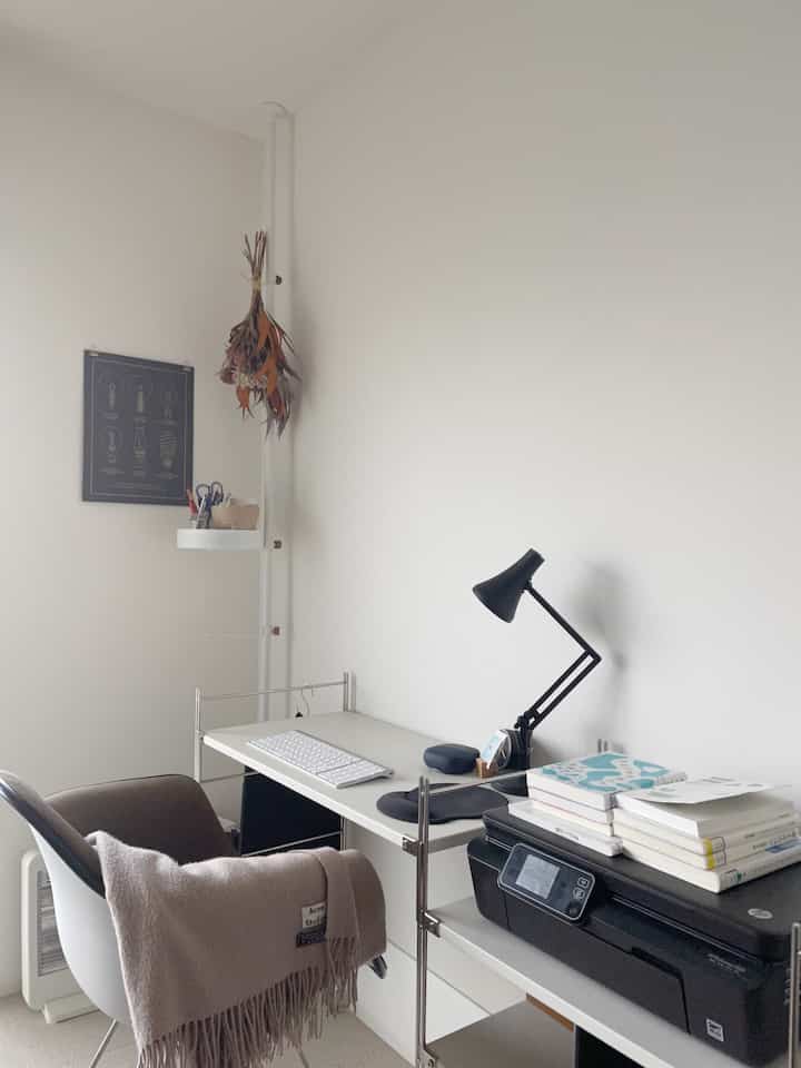 White-tone bedroom with a compact home office featuring a desk, black desk lamp, and organized books, creating a clean workspace