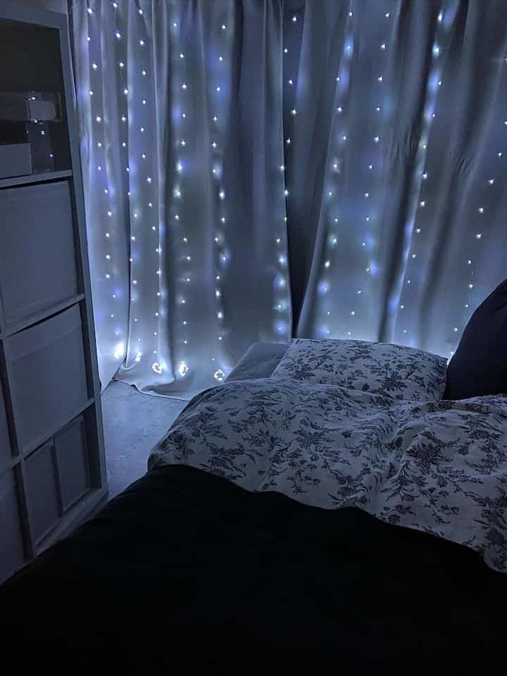 ホワイトトーンのカーテンと寝具がある寝室、LEDストリングライトが温かみのある雰囲気を演出している。