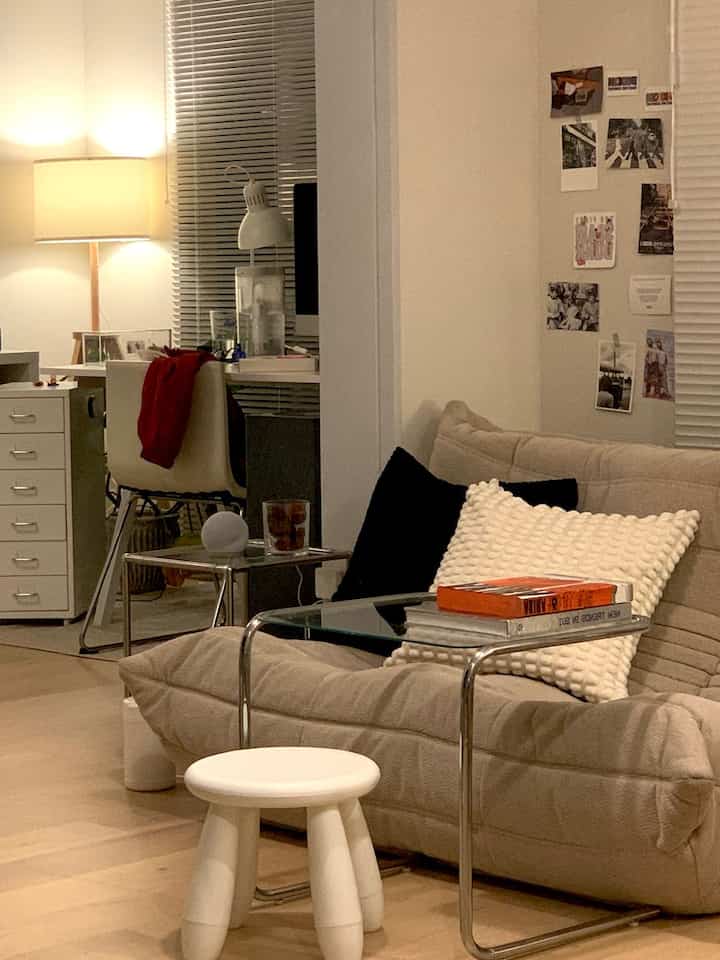 Beige-toned living room featuring a sofa, glass side table, and a desk with lamps reflected in a large mirror, creating a cozy ambiance