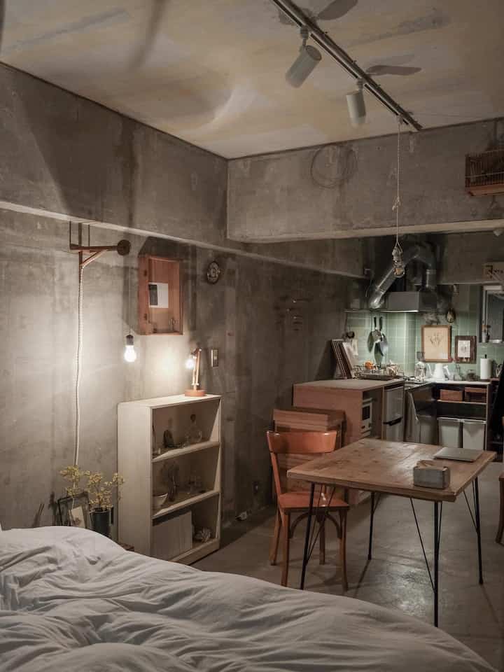 ナチュラルカラーとヴィンテージ感のあるワンルームの寝室とキッチン、シンプルな木製家具と露出したコンクリート壁が調和した雰囲気のあるインテリア