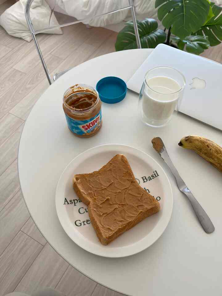 白とベージュのトーンでまとめられた丸いダイニングテーブルに、ピーナッツバターが塗られたトースト、バナナ、ミルクのグラスとノートパソコンが置かれた朝食のある雰囲気のインテリア