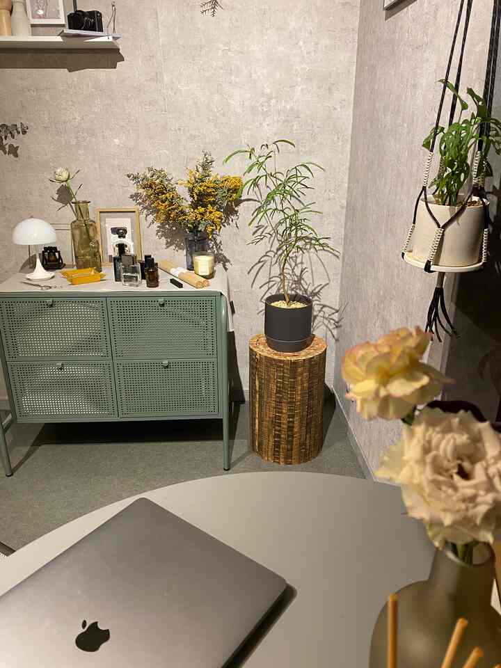 ナチュラルな色調のリビング、木製スツールの上に植物が置かれ、薄いグリーンのサイドボードと花瓶が配置された落ち着いた雰囲気のインテリア