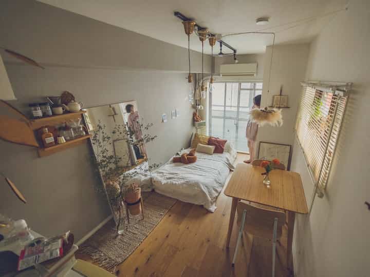 Natural tone compact and cozy studio apartment featuring bed, wooden dining table, wall-mounted shelf, and large mirror