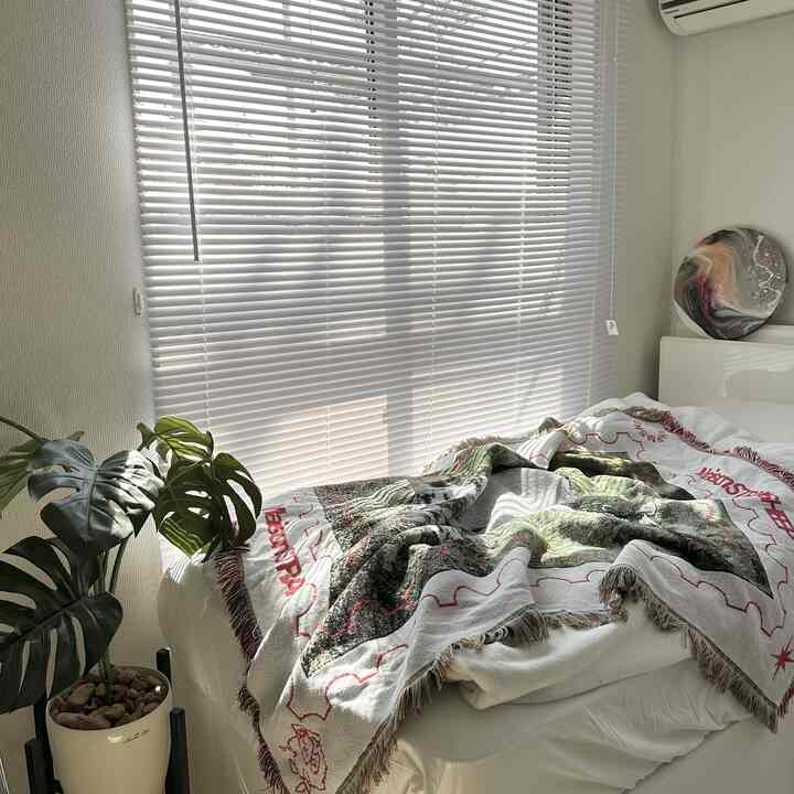 明るい日差しが差し込む白いブラインドの寝室、模様のある掛け布団カバーと観葉植物が配置された雰囲気のあるインテリア