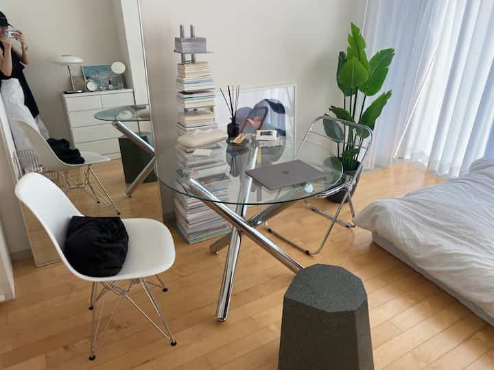 白と木目調の調和がとれたダイニング、ガラスの食卓と椅子、ベッド、植物が配置されたシンプルな空間