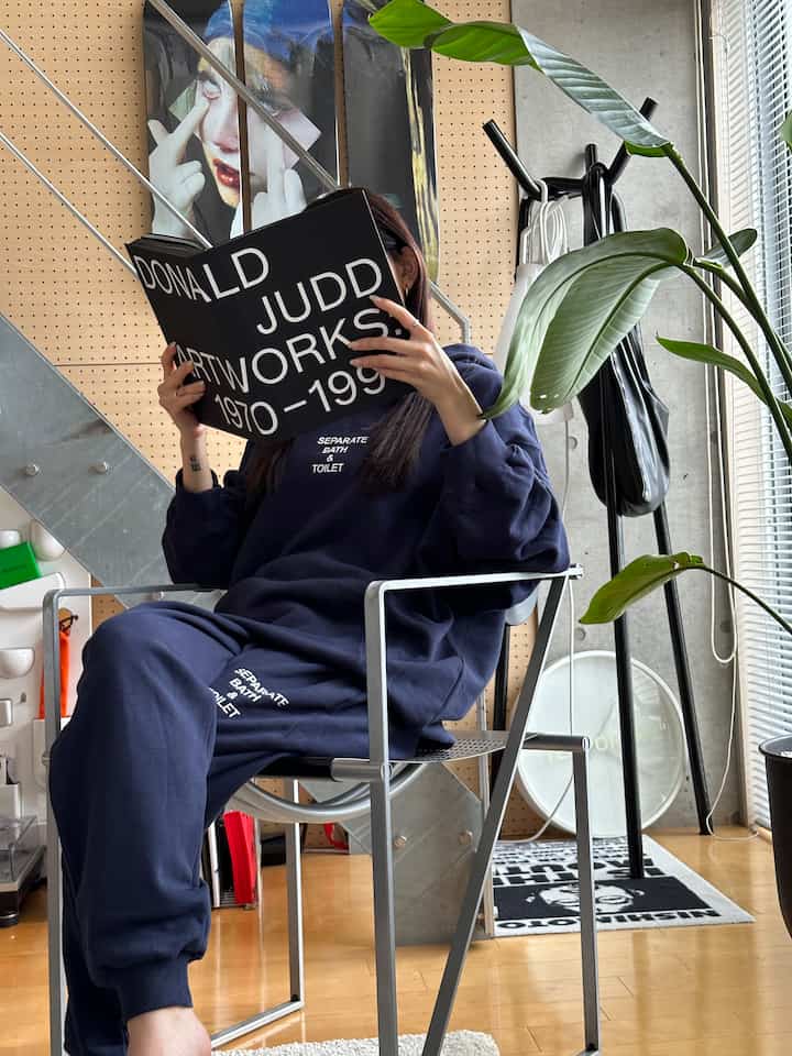 Modern industrial interior with gray concrete wall and wooden floor, featuring a person sitting on a black metal chair reading a large book and a green plant nearby