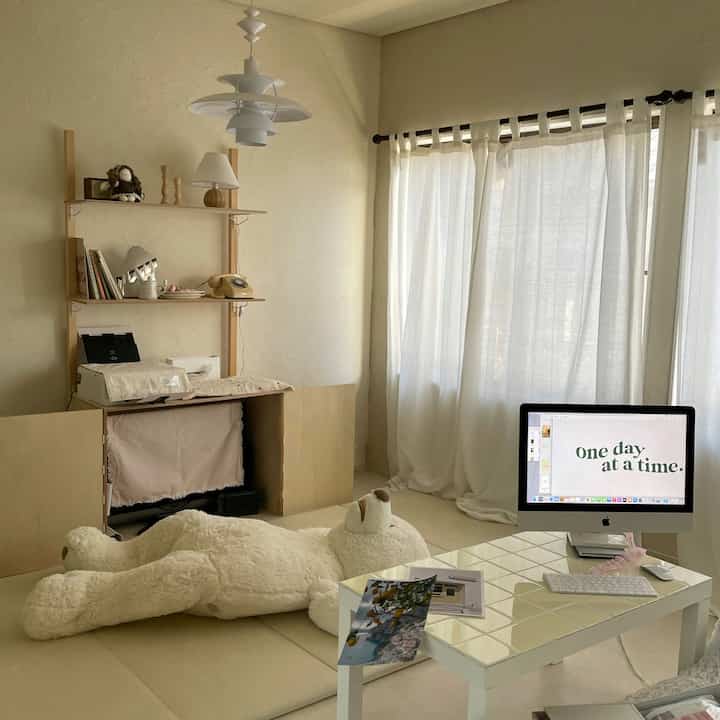 温かみのあるベージュと白の色調のリビング、巨大なクマのぬいぐるみとiMac、タイルのテーブルが配置された印象的なホームオフィス空間