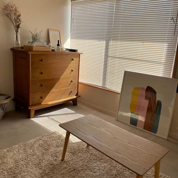 ベージュの床タイルとナチュラルな木製チェスト、白いブラインド、低めの木製テーブルが配置された居心地の良いリビング