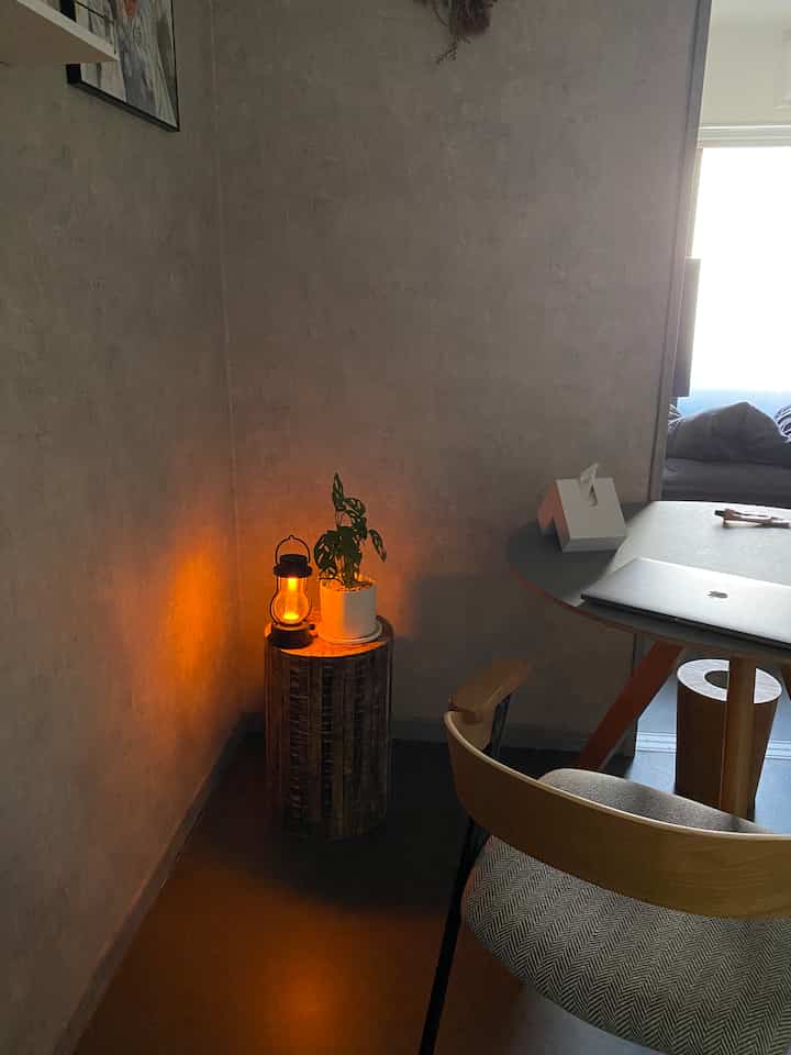 ナチュラルカラーの小さな部屋のコーナーに、木製スツールとバルミューダのランタン、植物が配置されたモダンなホームオフィス空間