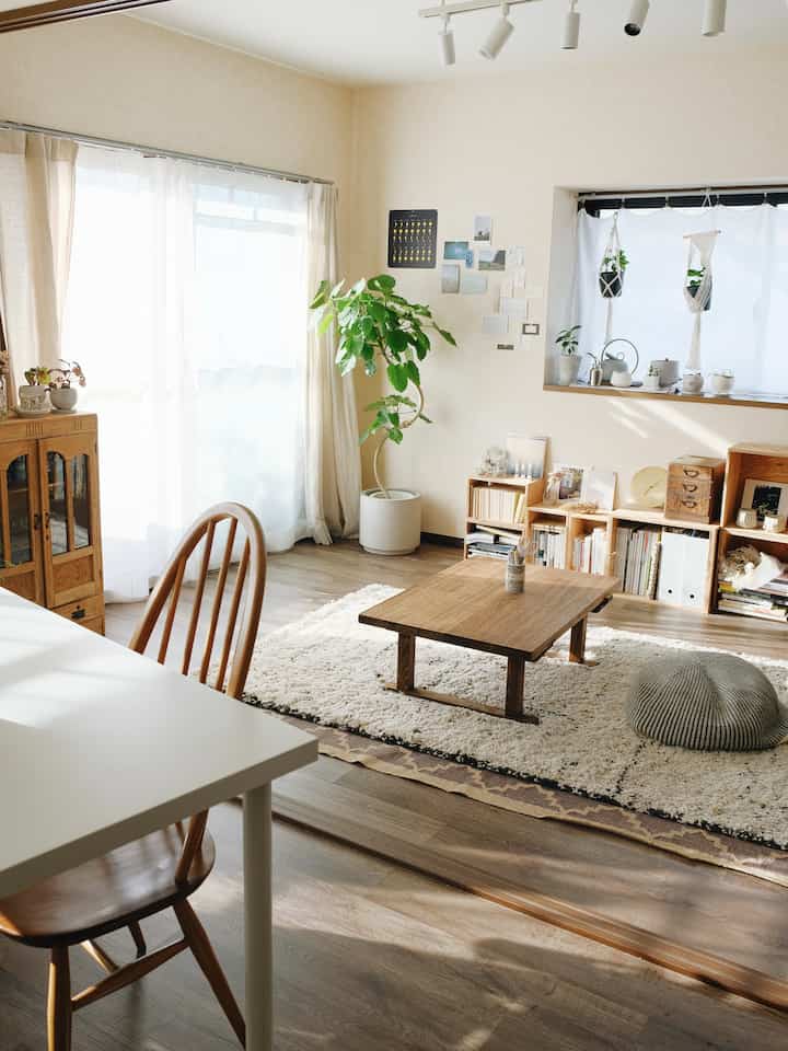 ナチュラルな木目調と白い壁の1LDKリビング、低い木製テーブルとクッション、多数の植物が配置された温かみのある空間