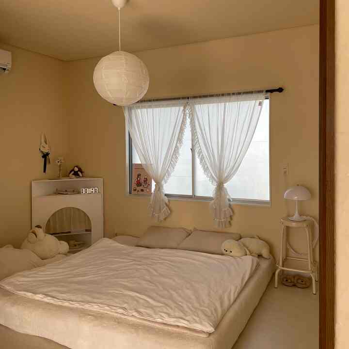 ベージュの色調が特徴的な寝室、大きな低いベッドと柔らかな自然光が入るカーテン付きの窓、かわいらしいぬいぐるみとスツールが並ぶ落ち着いたインテリア