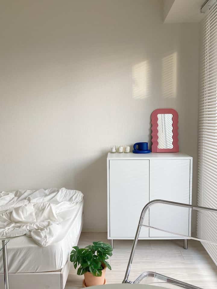 A white and pink toned bedroom featuring a cabinet with a pink wave mirror and a plant, creating a clean atmosphere