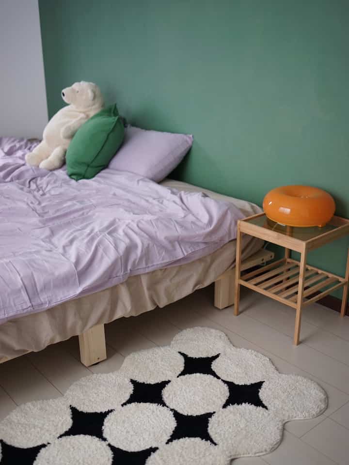 Bedroom with lavender bedding and green wall, featuring wooden bed and side table in a simple natural setting
