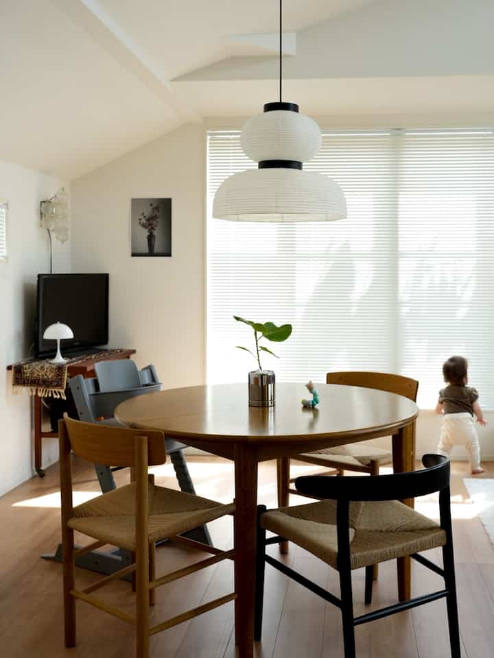 ライトブラウンと白の色調のダイニング、円形テーブルと椅子が配置され、窓辺に立つ幼児のいる雰囲気のあるインテリア