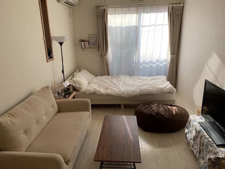 ベージュの自然な色調のワンルーム寝室、ソファとベッド、カーテン、フロアランプが配置された落ち着いたインテリア