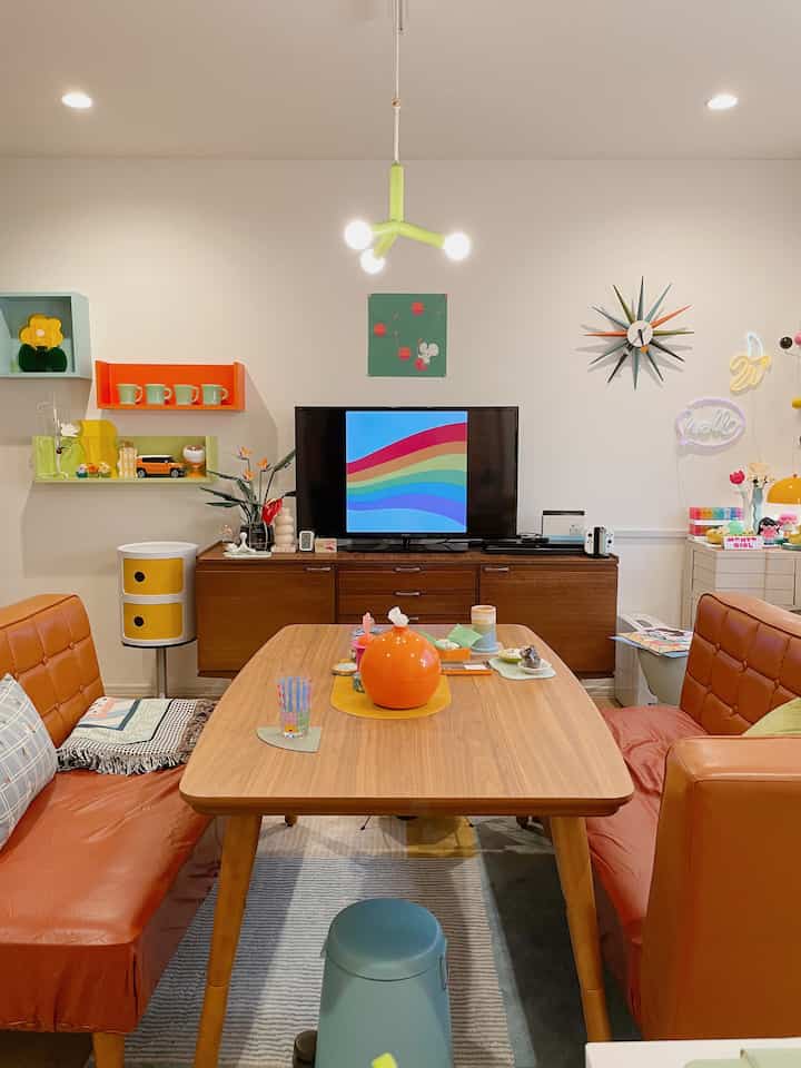 オレンジ色のレザーソファが配置されたミッドセンチュリーモダンスタイルの二人暮らしのリビング、カラフルな装飾と木製ダイニングテーブルがある温かみのある空間