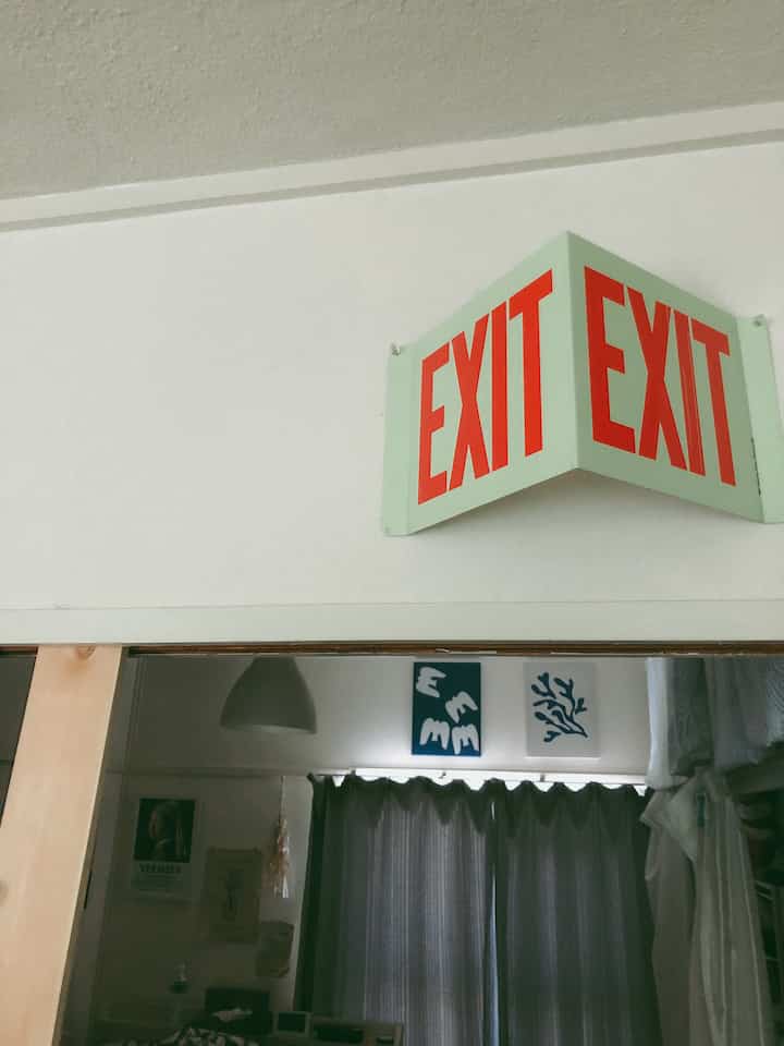 白い壁と赤いEXITサインがある玄関スペース、ダークなカーテンと壁アートが配置されたシンプルな空間