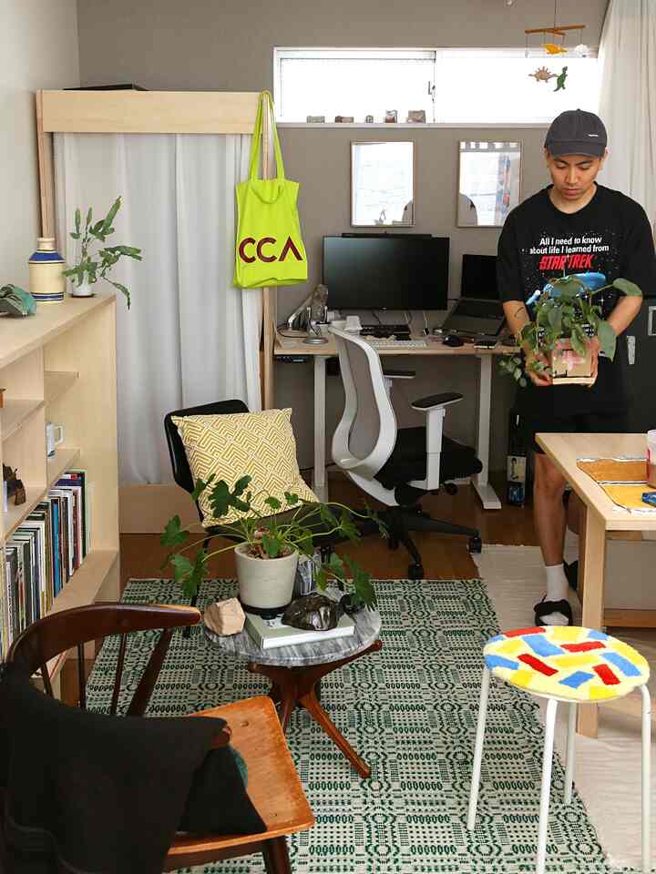 ナチュラルトーンと白いカーテンが調和する小さなホームオフィスに、複数の観葉植物と木製家具、カラフルなスツールが配置された雰囲気のあるインテリア