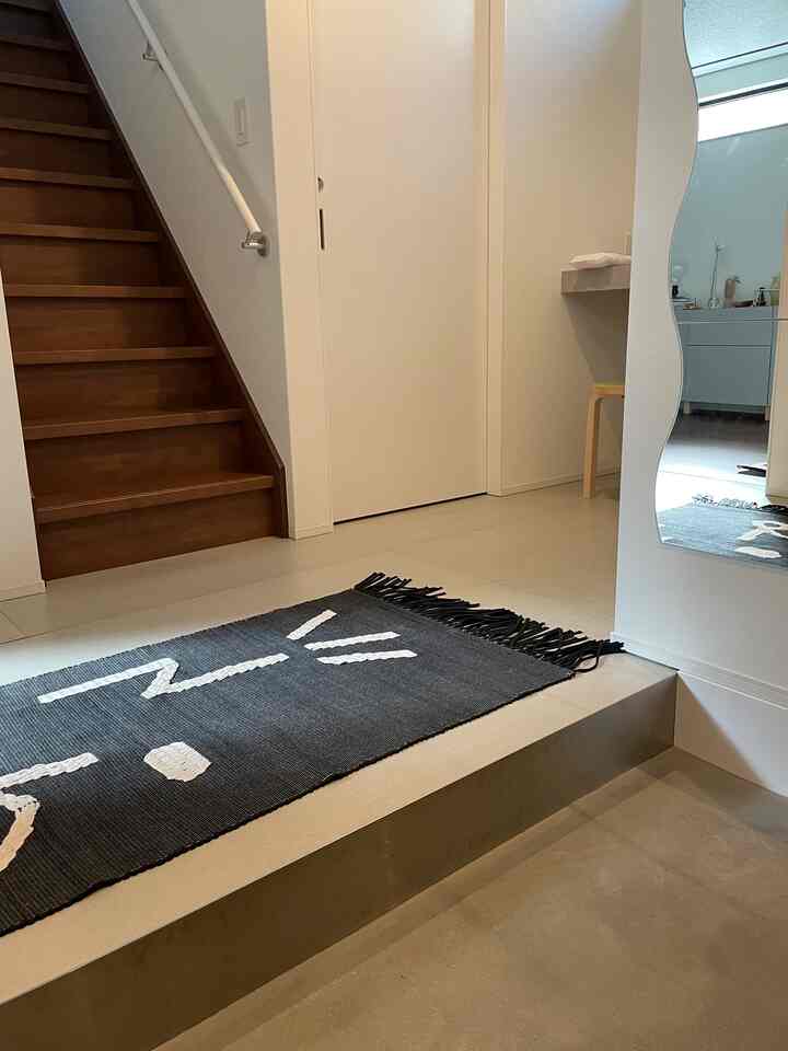 白い壁と木目調の階段があるシンプルなモダン玄関、黒の模様入りラグが配置された空間