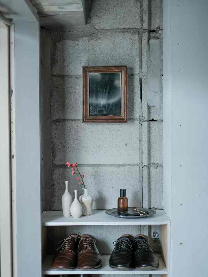 Cozy entrance space featuring gray concrete wall, white shelves with white vases, brown and black shoes, and a small framed mirror