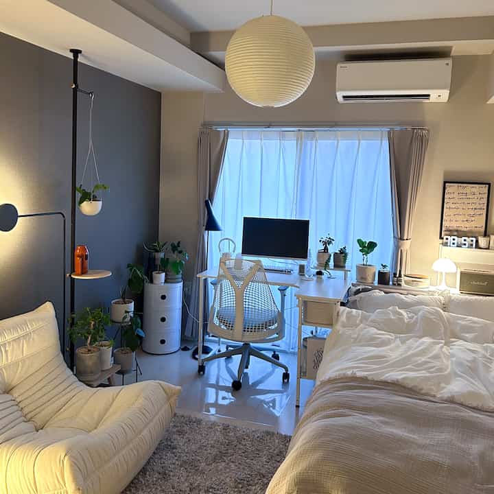 ホワイトとグレーの色調が調和するワンルーム、ベッドとデスク、植物が配置された落ち着いたシンプルなホームオフィスのあるインテリア