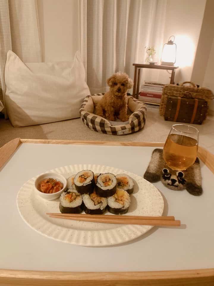 暖かい黄色の照明が灯るリビング、ホワイトのクッションソファとブラウンのペットベッドが配置され、トレイテーブルにキンパが置かれた雰囲気のあるインテリア