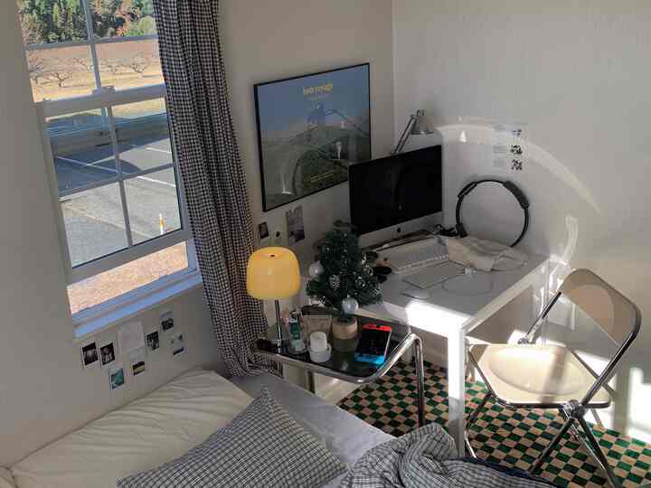 白い壁と家具が配置されたコンパクトな4.5畳の部屋に、緑のチェック柄ラグとレトロなテーブルランプが温かみのある雰囲気を演出する印象的なインテリア