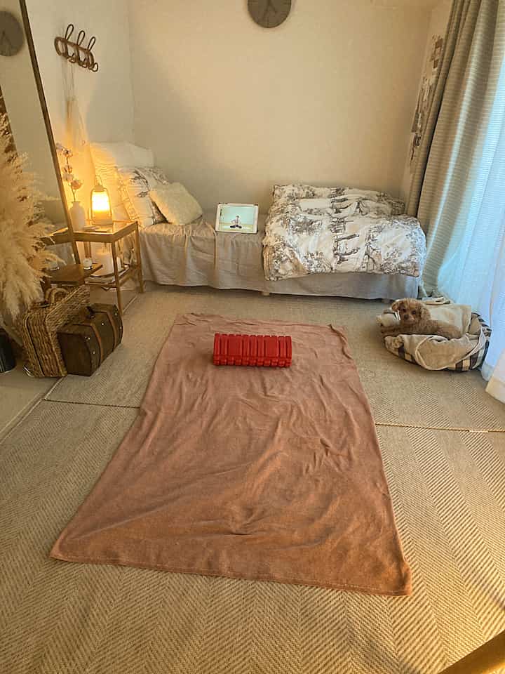 ベージュのナチュラルトーンのリビング、ベッドとナイトテーブル、大きなラグ、ペットの犬が配置された雰囲気のあるインテリア