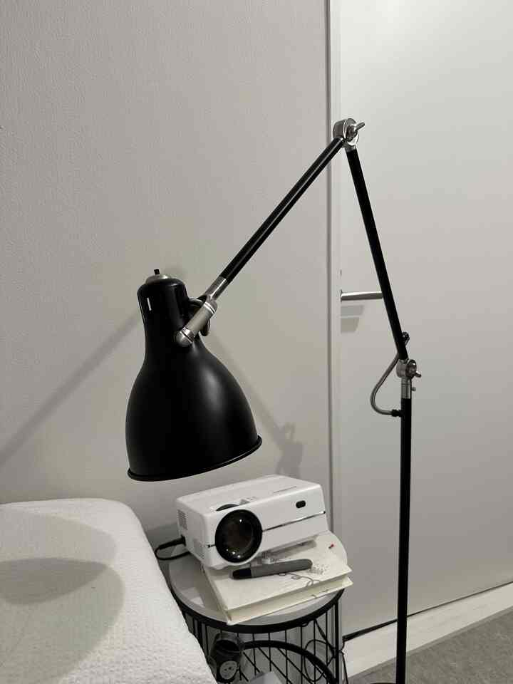 ホワイトとブラックのトーンが特徴の寝室、ミニマルな空間にサイドテーブルのプロジェクターとフロアランプが配置されている