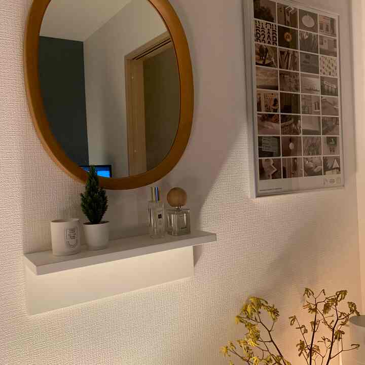 ナチュラルカラーとホワイトトーンの玄関の空間、丸い木製フレームの鏡と白い壁棚に小さな植物とキャンドルが置かれたシンプルなインテリア