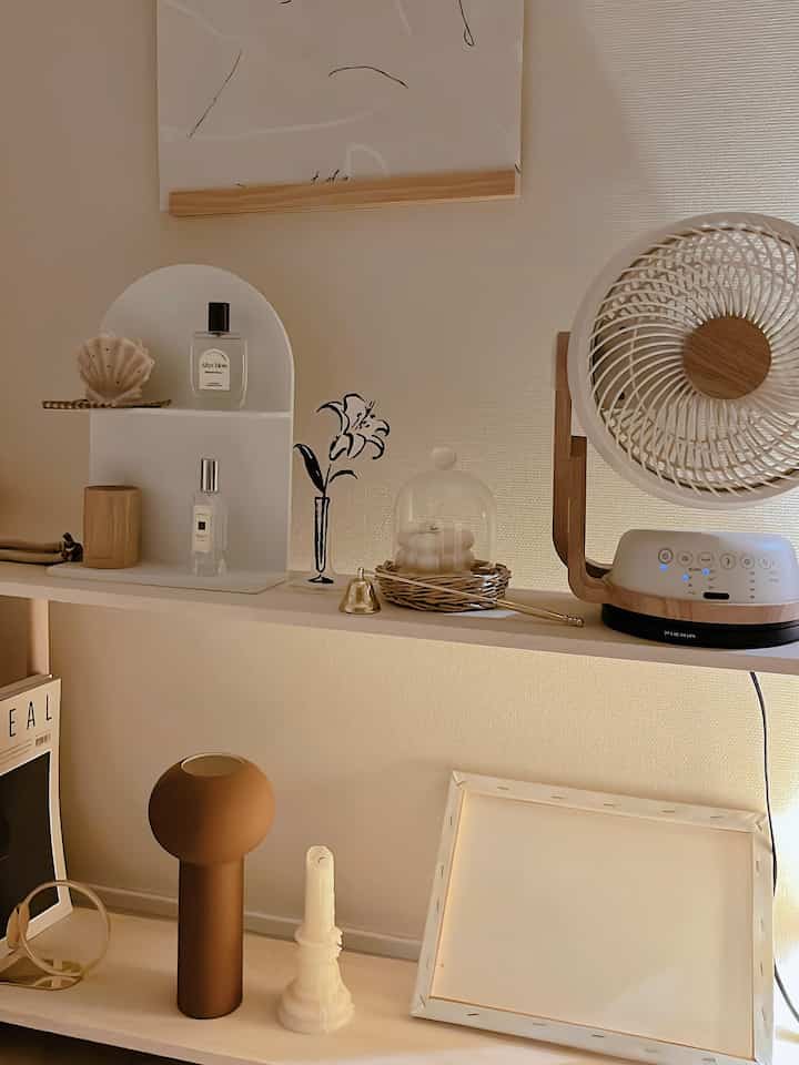 柔らかいベージュと白色調の寝室の棚、花瓶やキャンドル、扇風機などが配置された雰囲気のあるインテリア
