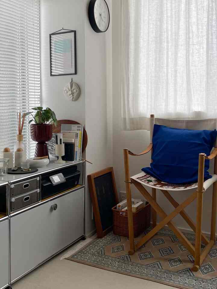 A white and natural tone living room corner featuring a wooden chair with blue cushion, storage cabinet, and decorative objects in a simple, calm atmosphere