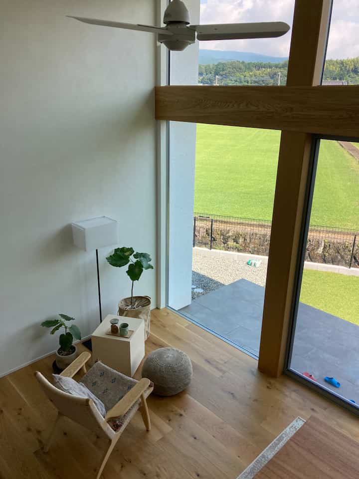 Natural tone room featuring wooden floor, armchair, stool, floor lamp, and plants beside a balcony with large windows