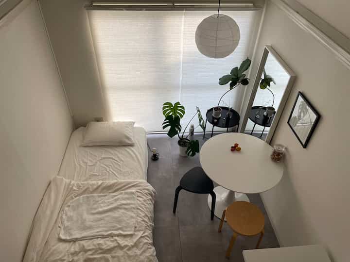 Bright white-toned small room featuring a single bed, round dining table, stools, and plants with minimalist atmosphere