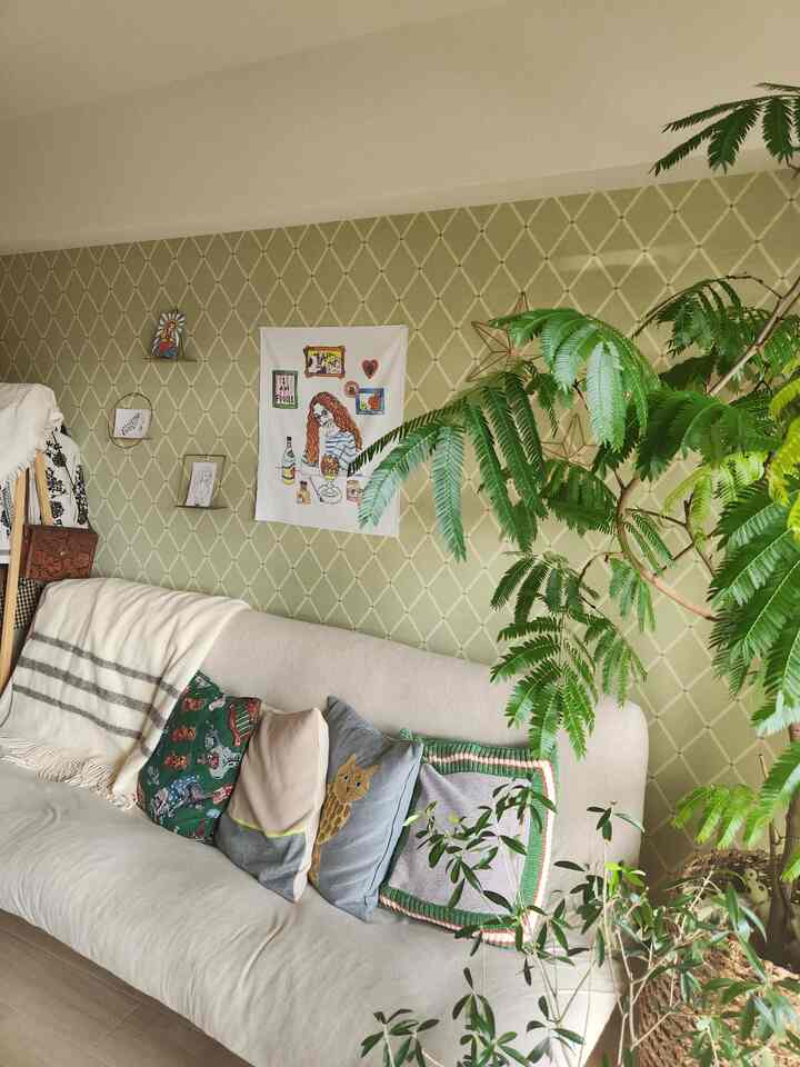 淡い緑色の壁紙とベージュのソファ、様々なクッションと大きな観葉植物が配置された自然な雰囲気のリビング