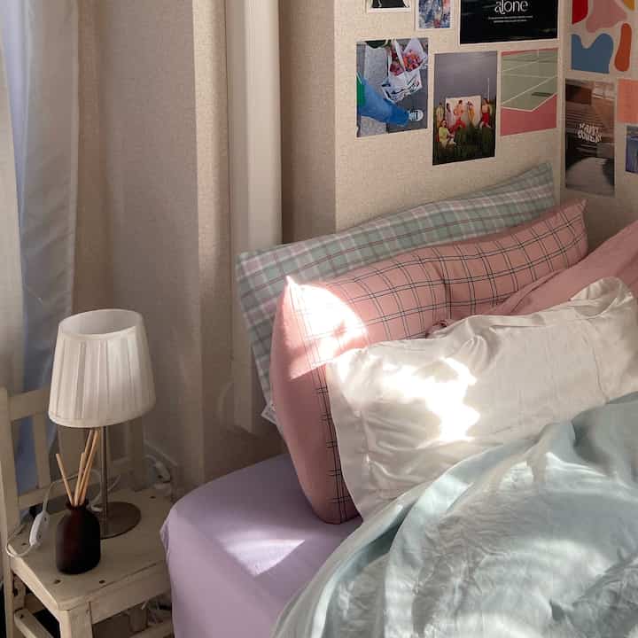 ナチュラルカラーの寝室、ピンクチェックの枕と白い枕が置かれたベッドとテーブルランプが配置された落ち着いたインテリア