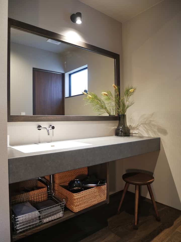 ナチュラルな色調の化粧台空間。大きな鏡、ラタンバスケット、木製スツールが配置された落ち着いたバスルームの雰囲気