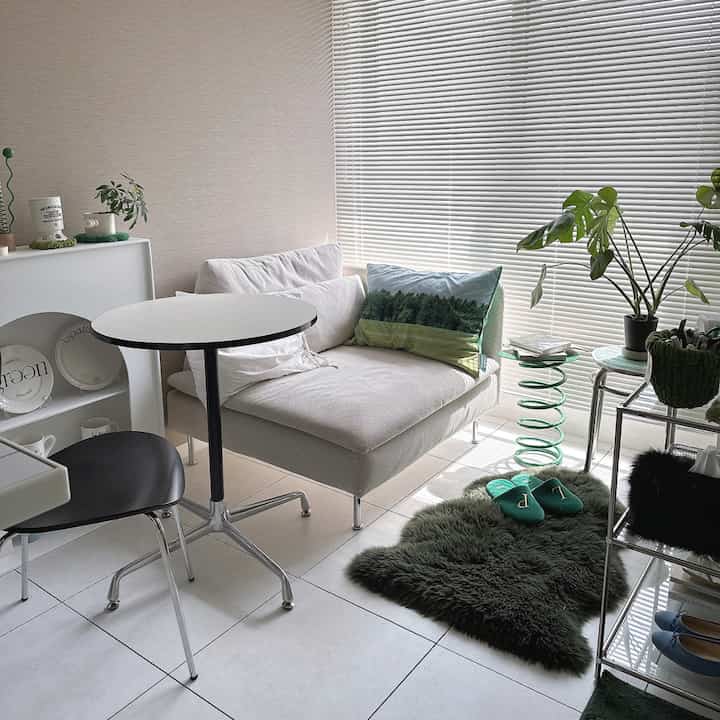 白と緑のトーンのリビング、白いコーナーソファと緑のラグ、植物が配置された自然で落ち着いたインテリア