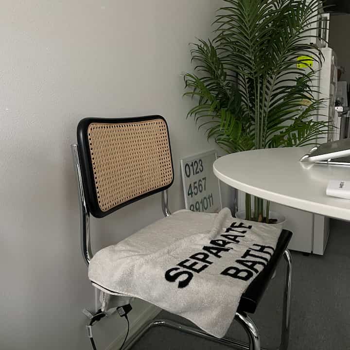 Compact home office space with gray floor and white wall, featuring black frame armchair with natural cane backrest and a white towel draped on it