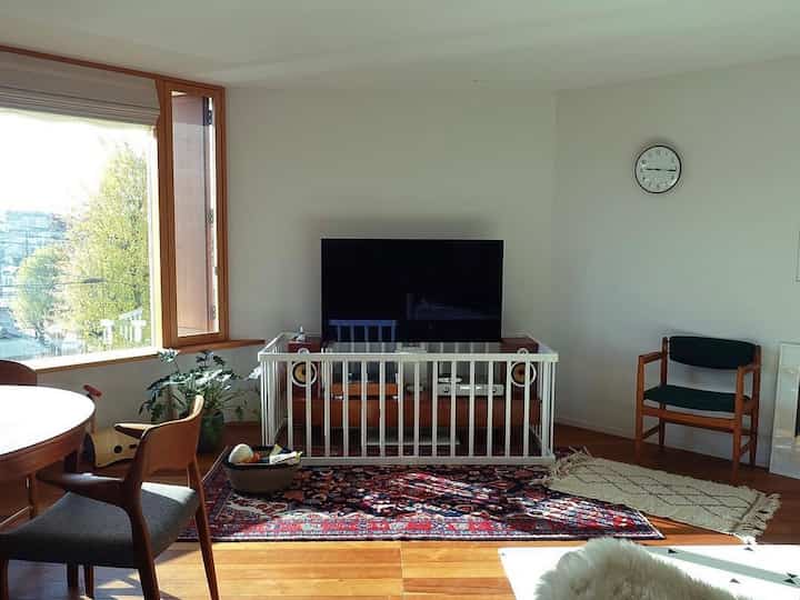 White-walled living room with wood-toned furniture, featuring a central baby safety gate in front of the TV and dining chairs, creating a warm atmosphere