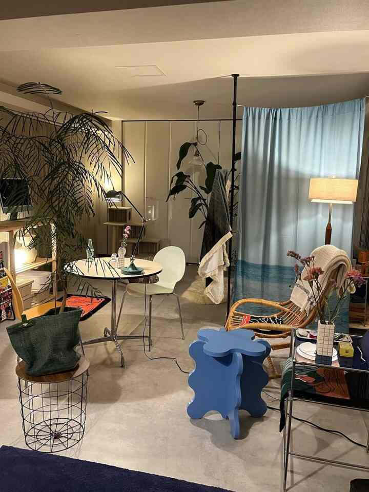 ナチュラルな色調のリビング、右側にブルーのカーテンとフロアランプ、中央に丸いダイニングテーブルと椅子が配置された印象的な空間