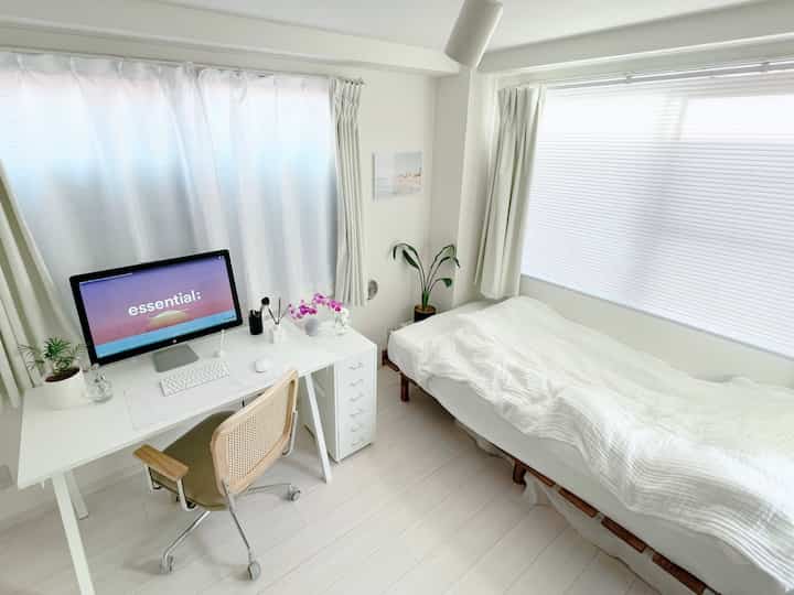 明るくすっきりした白を基調とした狭い寝室、白いデスクとベッド、小さな植物が配置された清潔感あるホームオフィスのインテリア