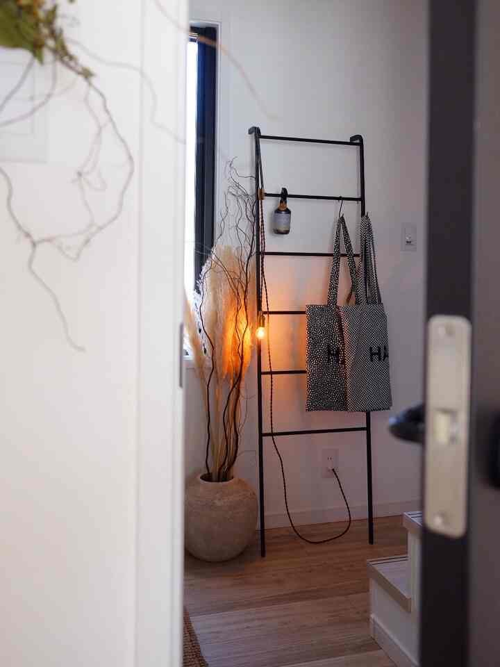 ナチュラルカラーと木目調の調和が美しい玄関、黒い金属製ラダーにエコバッグが掛けられ、温かみのある照明が配置された印象的な空間