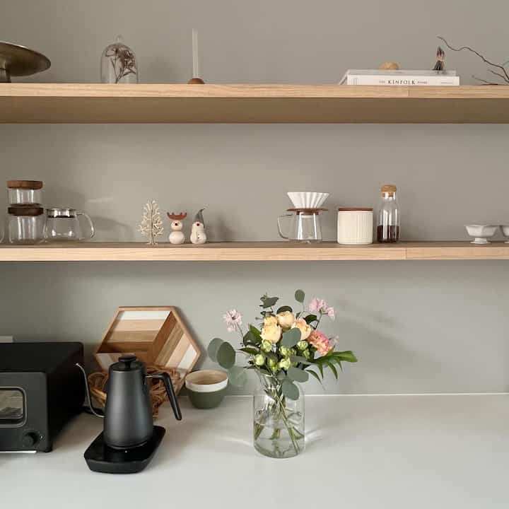 ナチュラルなホワイトトーンのキッチン、木製の棚と透明なガラス製品、花瓶に生けられた花がある清潔感のある空間
