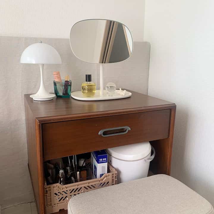 ナチュラルなベージュとブラウンの色調のドレッサースペース、ヴィンテージの木製チェスト、楕円形の鏡、白いテーブルランプが配置された雰囲気のあるインテリア