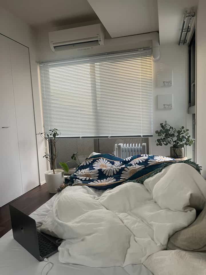 白と茶色のトーンの寝室、大きな窓にブラインドが設置され、植物が配置された落ち着いたモダンな空間