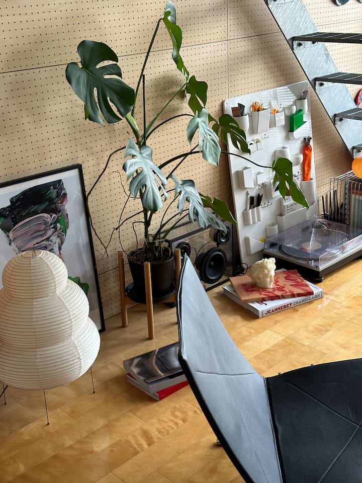 Modern space with wood-tone flooring and beige pegboard wall, featuring a large Monstera plant, white floor lamp, record player and decorative objets