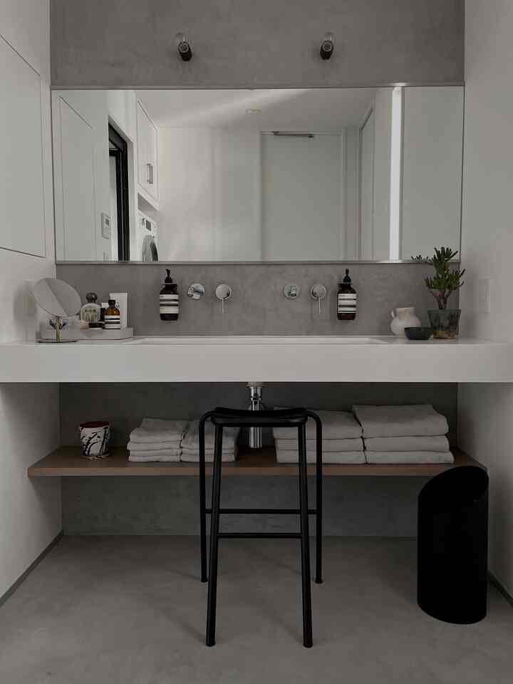 グレーと白のトーンのモダンなバスルームの洗面台空間で、タオルとスツールが配された清潔感のある無機質インテリア