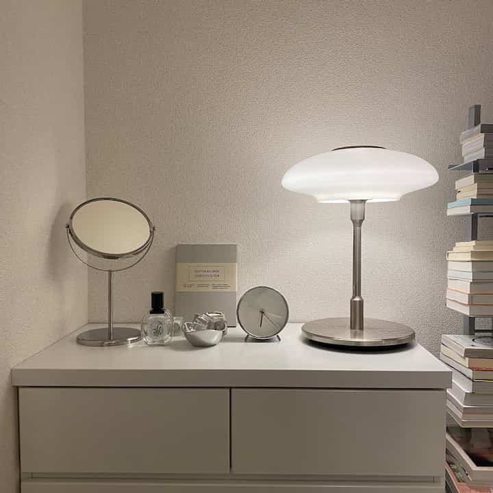 White and silver toned bedroom dresser with table lamp, round mirror, clock, and diffuser arranged neatly for clean interior