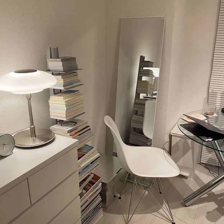 White-toned small corner room featuring a standing mirror, bookshelf, chair, and table lamp with a simple interior design